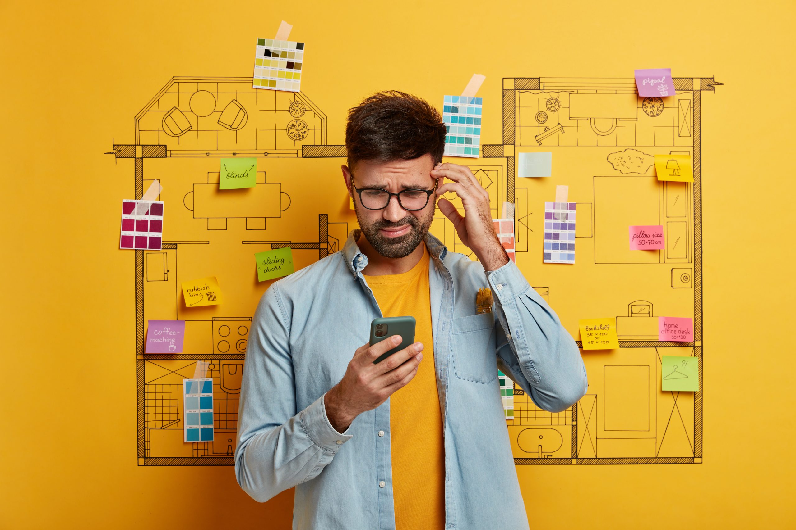 Puzzled bearded architect works with blueprint, works on architectural plan, focused in smartphone device with negative expression, stands against house design project, cannot find creative ideas