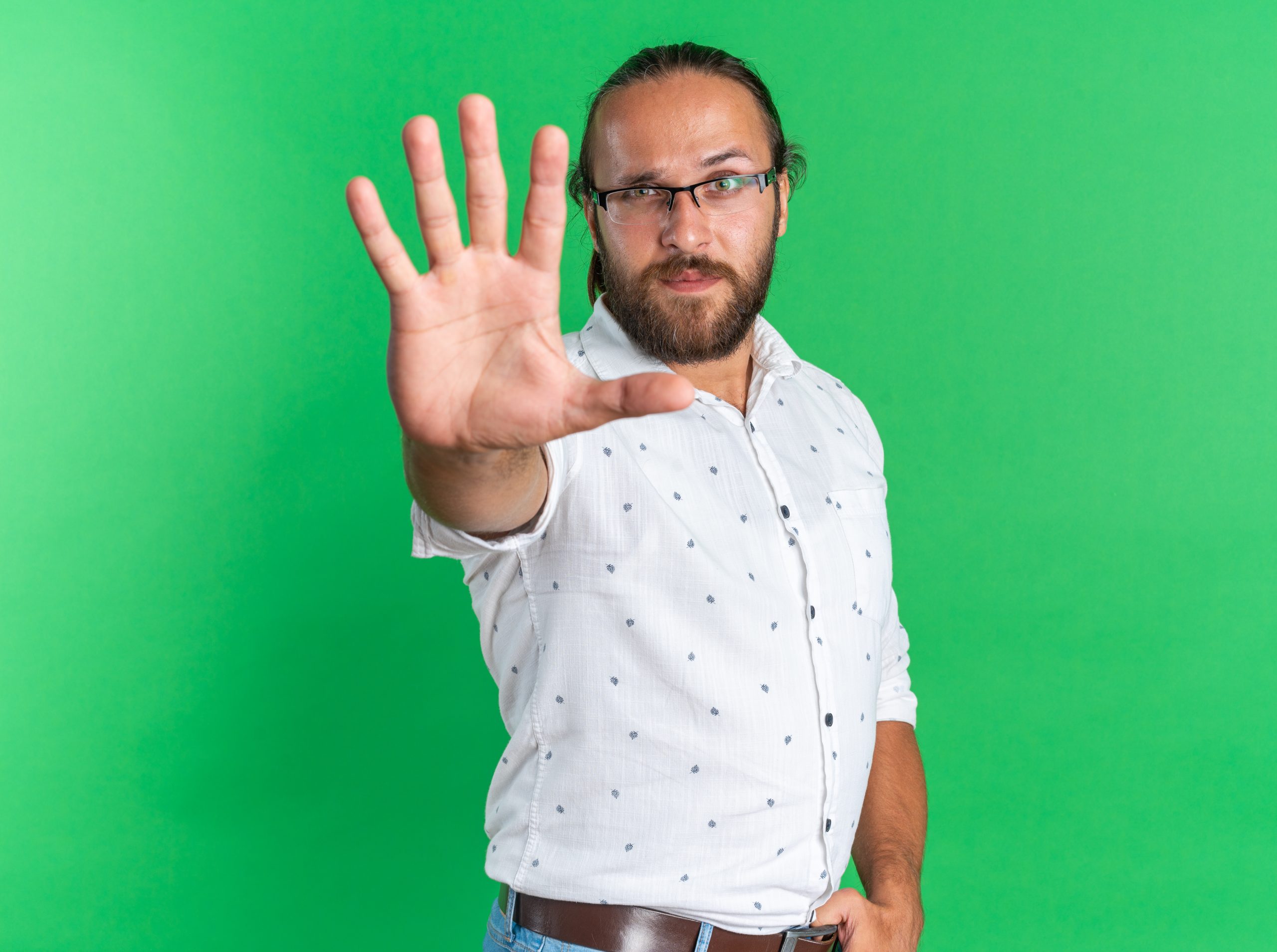 strict adult handsome man wearing glasses standing in profile view looking at camera keeping thumb in belt doing stop gesture isolated on green background with copy space